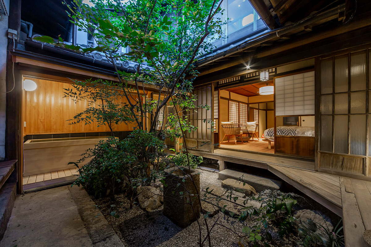 A traditional Japanese ryokan or inn, located in Tomonoura, Japan. The ryokan is a two-story wooden structure with a sloping roof and paper lanterns hanging outside. The interior of the ryokan is visible through the open sliding doors, revealing traditional tatami flooring, sliding shoji doors, and futon beds laid out on the floor. The image conveys a sense of tranquility, simplicity, and harmony with nature, which is characteristic of the traditional Japanese aesthetic.